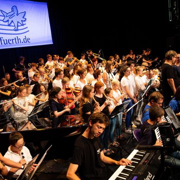 Bühne voller Menschen unterschiedlichen Alters mit verschiedenen Instrumenten, im Hintergrund ein Banner mit dem Logo der Musikschule