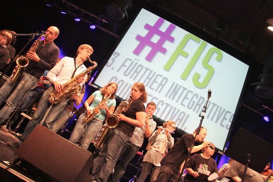 Viele Menschen mit Saxofon auf der Bühne, im Hintergrund Videoleonwand mit dem Logo des 3. Fürther Integrativen Soundfestivals