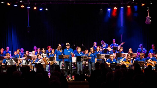 Vollgas Connected: rund 30 Musiker:innen im Festival-T-Shirt stehen und sitzen im Scheinwerferlicht auf der Bühne