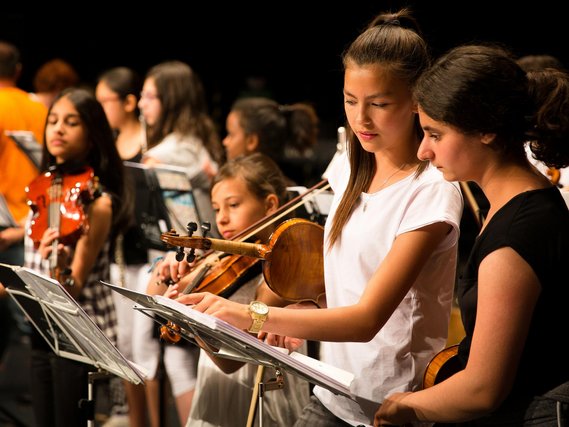 Zwei Mädchen mit Violinen treffen letzte Absprachen kurz vorm Auftritt, im Hintergrund sind weitere jugendliche Orchestermitglieder zu sehen.