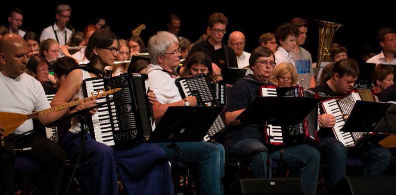Mehrere Frauen und ein Junge mit Down-Syndrom spielen gemeinsam Akkordeon im Vordergrund des großen Orchesters. Am linken Bildrand spielt ein Mann eine orientalische Langhalslaute
