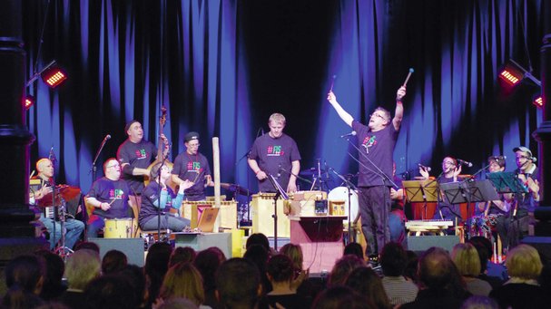Ensemble mit verschiedenen Orff-Instrumenten, in der Mitte ein Mann, der beide Arme nach oben streckt, alle tragen das Festival-T-Shirt FIS 2013