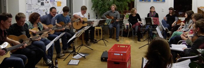 Jugendliche und Erwachsene sitzen im Kreis, vor sich Notenständer, einige spielen Gitarre, andere singen