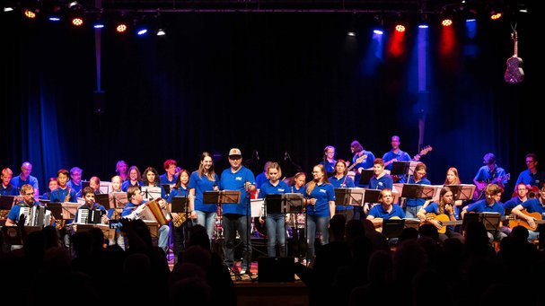 Vollgas Connected: rund 30 Musiker:innen im Festival-T-Shirt stehen und sitzen im Scheinwerferlicht auf der Bühne