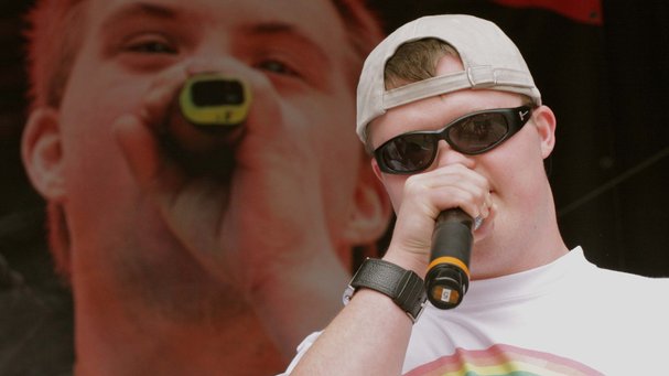 Ein junger Mann mit Sonnenbrille, in der Hand ein Mikro. Im Hintergrund eine große Videoleinwand