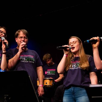 ein Sänger und drei Sängerinnen mit Mikros, alle tragen das Festival-T-Shirt FIS 2019