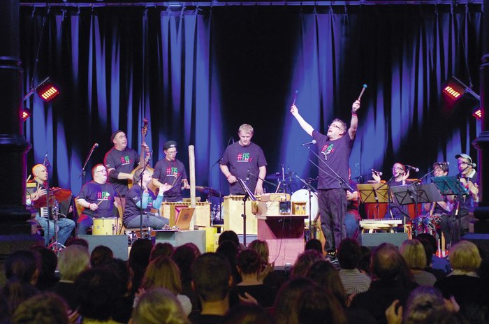 Ensemble mit verschiedenen Orff-Instrumenten. In der Mitte ein Mann, der beide Arme nach oben streckt. Alle tragen das Festival-T-Shirt FIS 2013