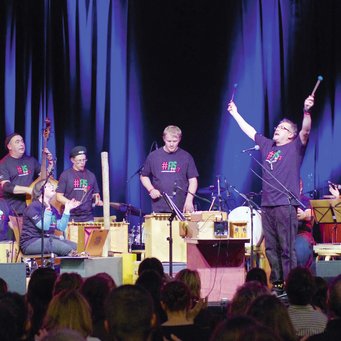 Ensemble mit verschiedenen Orff-Instrumenten, in der Mitte ein Mann, der beide Arme nach oben streckt, alle tragen das Festival-T-Shirt FIS 2013