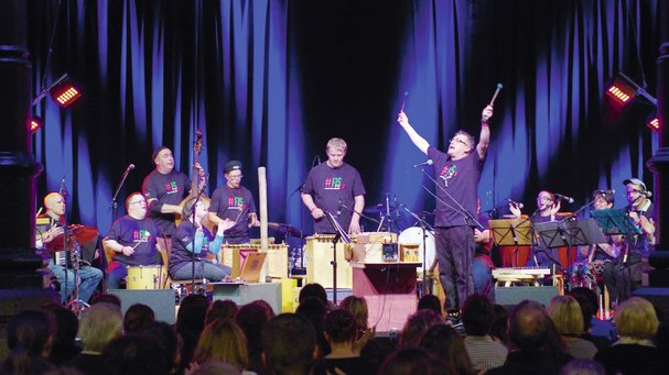 Ensemble mit verschiedenen Orff-Instrumenten, in der Mitte ein Mann, der beide Arme nach oben streckt, alle tragen das Festival-T-Shirt FIS 2013
