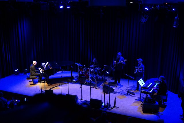 Klavierdozent Dieter Köhnlein am Flügel im Scheinwerferlicht. Im blaueen dunklen Hintergrund die Band.