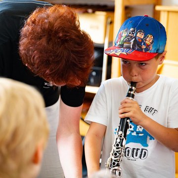 Kind mit Käppi bläst in eine Klarinette, die Lehrerin beugt sich helfend hinunter