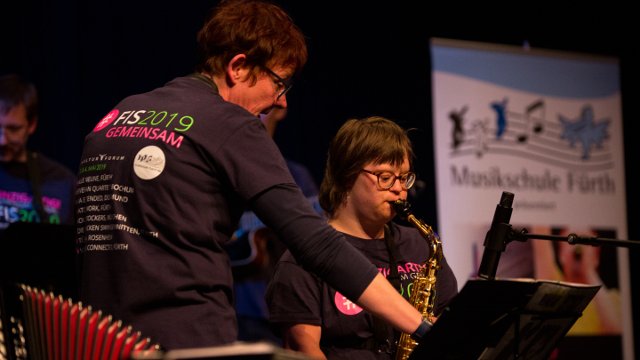 Musikerin mit Down-Syndrom spielt Saxophon, die Lehrerin deutet helfend in die Noten