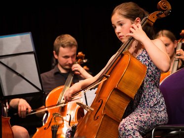 Mädchen spielt Cello, im Hintergrund weitere Cellist:innen