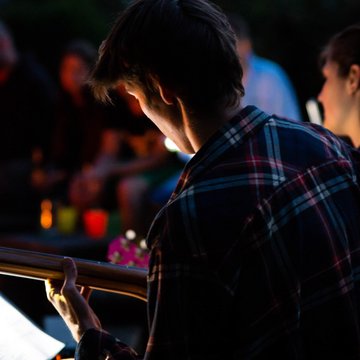 Junger Mann sitzt mit Gitarre am Lagerfeuer, die Kamera schaut ihm von hinten über die Schulter