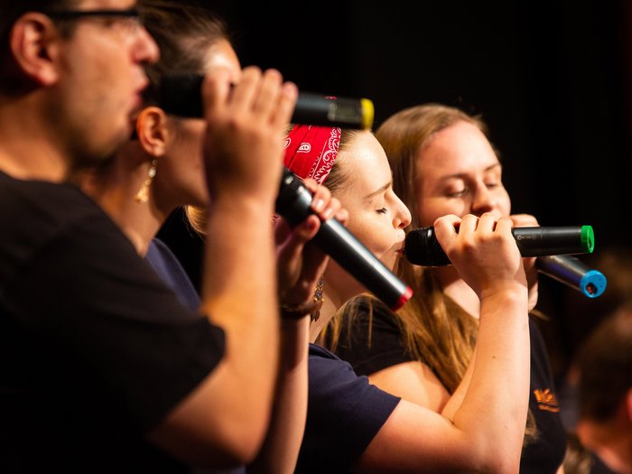 Vier Sänger:innen mit bunten Funkmikros. Die Sängerin im Zentrum trägt ein rotes Haarband und hat die Augen geschlossenen.