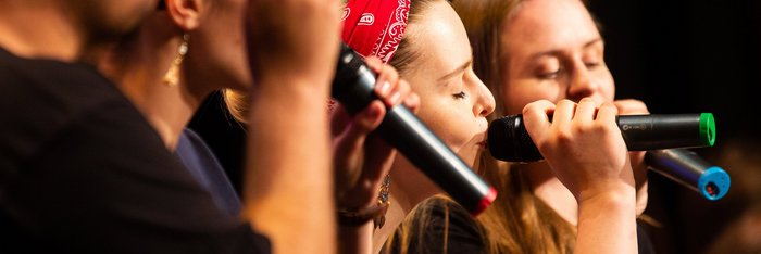 Vier Sänger:innen mit bunten Funkmikros. Die Sängerin im Zentrum trägt ein rotes Haarband und hat die Augen geschlossenen.
