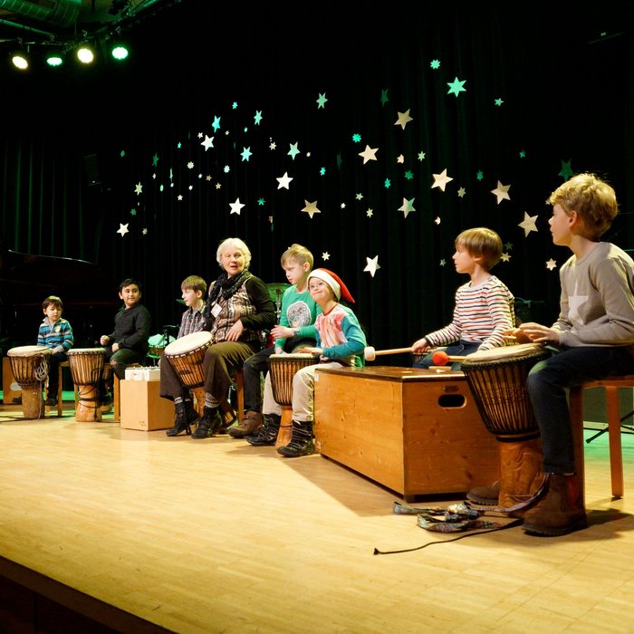 Kinder und Lehrerin spielen auf verschiedenen afrikanischen Trommeln, Weihnachtdeko im Hintergrund