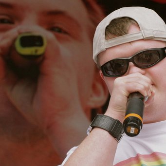 Ein junger Mann mit Sonnenbrille, in der Hand ein Mikro. Im Hintergrund eine große Videoleinwand