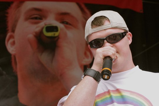 Junger Mann mit Sonnenbrille und Mikro, im Hintergrund große Videoleinwand