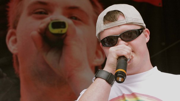Ein junger Mann mit Sonnenbrille, in der Hand ein Mikro. Im Hintergrund eine große Videoleinwand