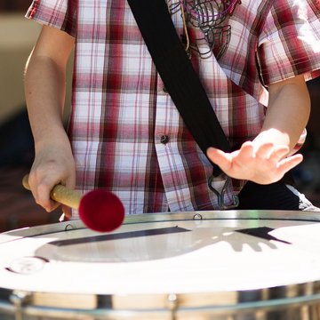 Oberkörper eines Kindes, quer über die Schulter ist eine große Sambatrommel geschnallt, in der rechten Hand einen dicken Schlägel