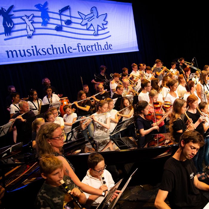 Große Bühne voller Schüler:innen. Kinder, Jugendliche, Erwachsene und Senior:innen musizieren gemeinsam. Im Vordergrund spielt Junge mit Down-Syndrom Blockflöte.