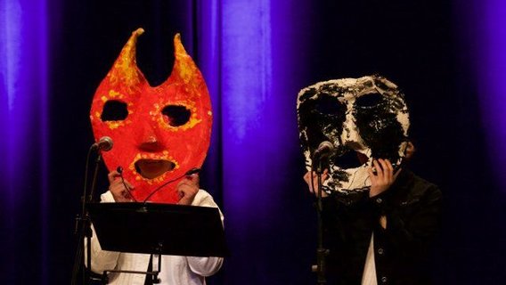 Zwei Menschen auf der Bühne, sie tragen kunstvolle große Masken