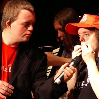 2 Menschen mit Behinderung. Während der eine Mundharmonnika spilet, hält ihm der andere das Mikro