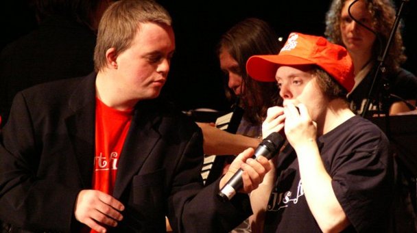 2 Menschen mit Behinderung. Während der eine Mundharmonnika spilet, hält ihm der andere das Mikro