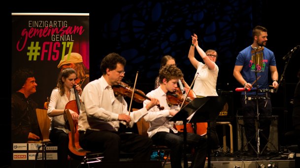 Das Ensemble piano plus mit Streichinstrumenten und eine Tänzerin. Im Hintegrund das Plakat des FIS 2017 mit dem Schriftzug Einzigartig. Gemeinsam genial.