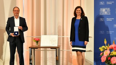 Robert Wagner präsentiert die soeben erhaltene Medaille. Daneben Landtagspräsidentin Ilse Aigner vor dem Logo des Bayerischen Landtages.