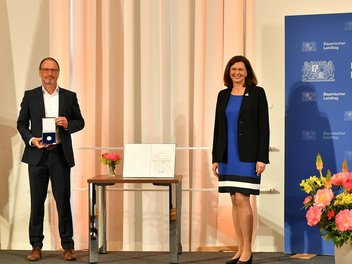 Robert Wagner präsentiert die soeben erhaltene Medaille. Daneben Landtagspräsidentin Ilse Aigner vor dem Logo des Bayerischen Landtages.
