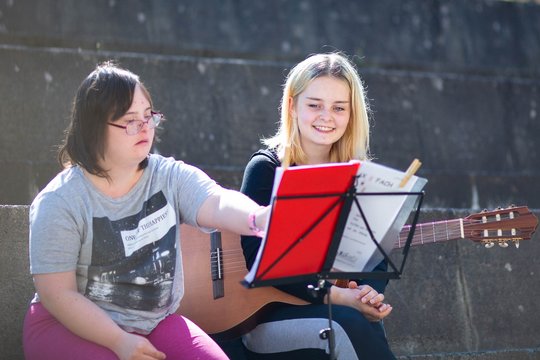 Zwei junge Frauen, eine davon mit Dosn-Syndorm sitzen im Sonnenschein vor einem Notenständer. Die eine spielt Gitarre, die andere deutet auf die Noten.