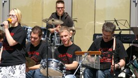 Musiker:innen mit Behinderung auf der Open Air Bühne. Im Vordergrund eine Sängerin, dahinter mehrere Menschen mit diversen Percussioninstrumenten und Schlagzeugestival der Musikschule Fürth