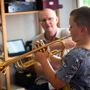Junge im Grundschulalter spielt Trompete, im Hintergrund unscharf der Lehrer