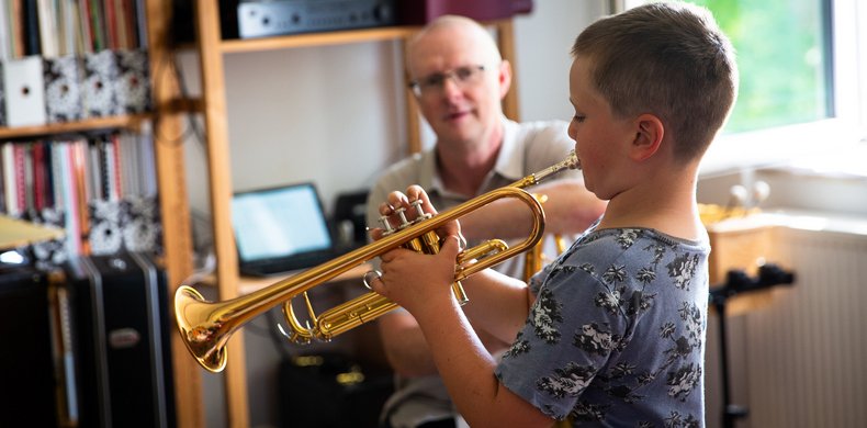 Junge im Grundschulalter spielt Trompete, im Hintergrund unscharf der Lehrer