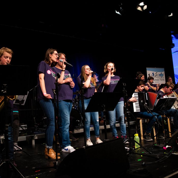 Eine Bühne voller Musiker:innen mit unterschiedlichen Instrumenten, im Zentrum ein Sänger und 3 Sängerinnen 