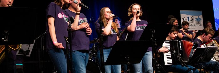 Eine Bühne voller Musiker:innen mit unterschiedlichen Instrumenten, im Zentrum ein Sänger und 3 Sängerinnen 
