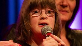 Mädchen singt auf der Bühne, eine Frau legt ihr einen Arm um die Schulter