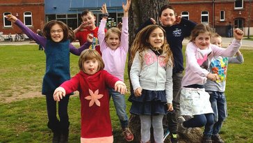 Kinder im Grundschulalter hüpfen auf der Wiese vor der Musikschule in die Höhe und strecken ihre Arme nach oben