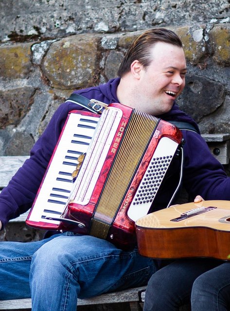 Akkordeonist mit Down-Syndrom und Gitarristin sitzen lachend auf einer Bank im Freien