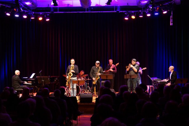 Die Saxofondozenten Norbert Emminger und Tobias Schöpker und Trompetendozent Stefan Schalanda in der ersten Reihe auf der Bühne. Hinter ihnen die Band. Im Vordergrund des Bildes sieht man die Rücken des Publikums.