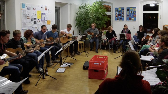 Jugendliche und Erwachsene sitzen im Kreis, vor sich Notenständer, einige spielen Gitarre, andere singen