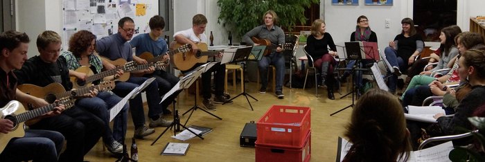 Jugendliche und Erwachsene sitzen im Kreis, vor sich Notenständer, einige spielen Gitarre, andere singen