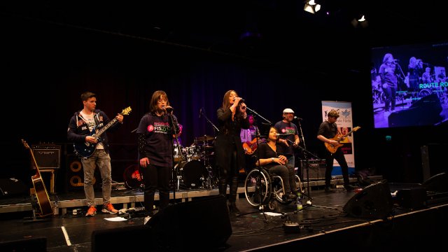 Die Band Route Rockers steht auf der Bühne, eine Sängerin sitzt im Rollstuhl