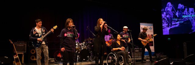 Die Band Route Rockers steht auf der Bühne, eine Sängerin sitzt im Rollstuhl