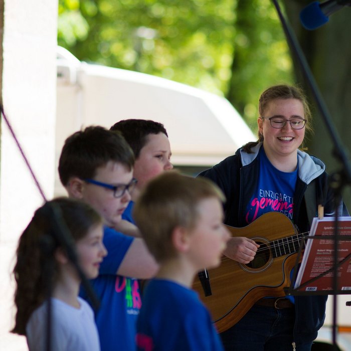 Kinder mit Behinderung singen bei einem Konzert im Freien, an der Gitarre von einer lachenden jungen Frau begleitet