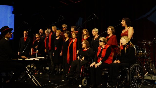 Der Heidelberger Beschwerdechor: sie tragen schwarz/rote Kleidung, in der ersten Reihe sitzen drei Sänger:innen im Rollstuhl