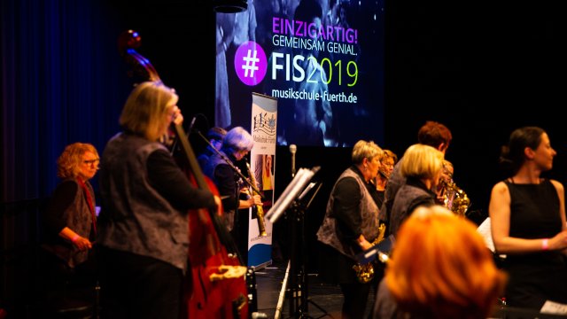 Seitliche Ansicht der Band Die schicken Swingschnitten, im  Hintergrund das Logo des FIS 2019 mit dem Schriftzug Einzigartig. Gemeinsam genial