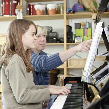 Mädchen sitzt lächelnd am Klavier, daneben ihre Lehrerin, die auf die Noten deutet